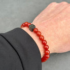 Close-up van een pols versierd met een Stoney Bracelets Red Onyx Stones (8mm) armband, met een opvallende zwarte kubusvormige bedel. Het individu is gekleed in een donker shirt met lange mouwen, tegen een grijze, neutrale achtergrond die de ingewikkelde kralen op de armband accentueert.