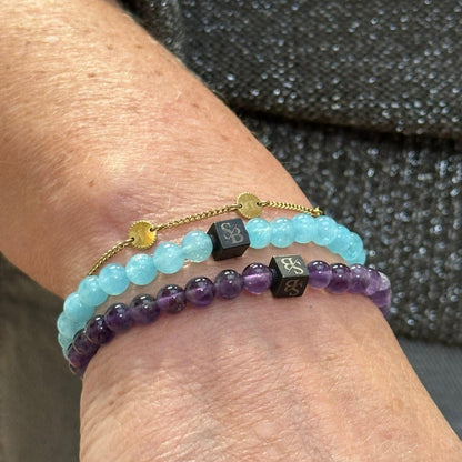 Een close-up van een pols versierd met twee kralenarmbanden (kralen armband) van Stoney Bracelets. Eén armband heeft lichtblauwe kralen met een kleine zwarte bedel, terwijl de andere is gemaakt met 6 mm Amethiststenen en een vergelijkbare zwarte bedel heeft. De persoon draagt een getextureerd grijs kledingstuk.