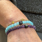 Een close-up van de pols van een persoon toont drie armbanden van Stoney Bracelets: een aquamarijn kralenarmband met het label "Aquamarine Stones (6mm)" versierd met een zwarte vierkante bedel, een gouden kettingarmband met kleine gouden kralen en een paarse kralenarmband ook met een zwarte vierkante bedel. Gemaakt van natuursteen en kralen, deze armbanden vormen een prachtige aanvulling op het grijze getextureerde shirt.