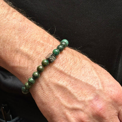 Een close-up van de pols van een man met een Stoney Bracelets' Dark African Turquoise Stones (8mm) kralenarmband, met een kleine zwarte vierkante kraal met een zilveren logo. De man heeft een lichte huidskleur en draagt een zwart shirt.