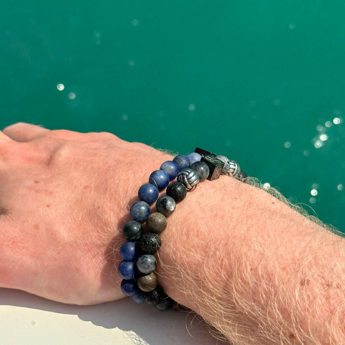 De pols van een persoon versierd met drie Stoney-armbanden, versierd met 8 mm blauwe Lapis Lazuli, grijze en zwarte natuurstenen, tegen een achtergrond van groen water.