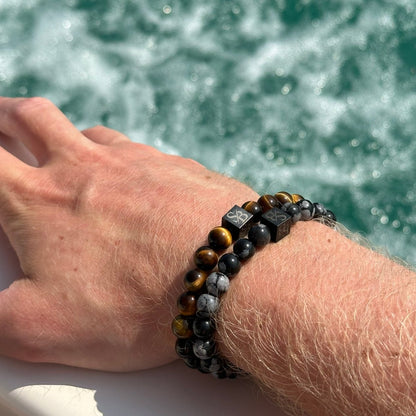 De hand van een persoon versierd met een Stoney Bracelets Gold Tiger Eye Stones (8mm) armband rust op een oppervlak in de buurt van water. De kralen zijn een mix van bruine tijgeroog en grijs gemarmerde ontwerpen. De achtergrond toont kolkend, schuimend water.