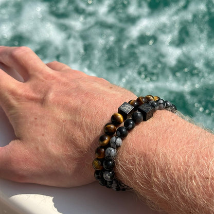 Een close-up van de pols van een persoon met twee Stoney Bracelets, een versierd met tijgeroog kralen en de andere met gemengde gekleurde edelstenen aangevuld met opvallende Snowflake Obsidian Stones (8mm) met witte symbolen. De achtergrond toont spattend wit water, waarschijnlijk afkomstig van het kielzog van een boot.