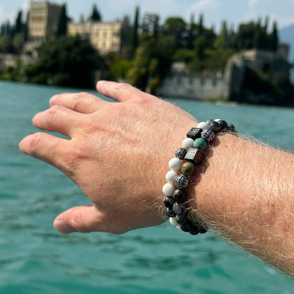 Een hand, versierd met twee kralenarmbanden en de elegante Hera | Silver+ Stones (8mm) natuursteen armband van Stoney Bracelets, strekt zich uit over een waterpartij. Op de achtergrond is een historisch gebouw te zien, omringd door bomen en groen. Het water is kalm en de lucht is helder.