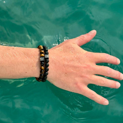 De hand van een persoon is gedeeltelijk ondergedompeld in groenblauw water. Ze dragen twee Stoney Bracelets armbanden. De ene heeft zwarte en bruine Lava Stones (8mm), terwijl de andere zwarte kralen heeft met kleine vierkante zilveren accenten.