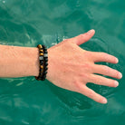 Een hand rustend op het wateroppervlak, versierd met twee Frosted Gold Tiger Eye Stones (8mm) kralenarmbanden van Stoney Bracelets, met metalen accenten en natuursteen details. De huid is licht vochtig, tegen een achtergrond van helder, groenachtig water.