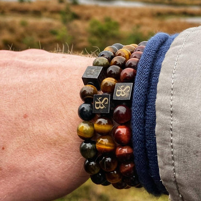 Een persoon versiert zijn pols met Stoney Bracelets' Threefold-Tiger Set (8mm), met verschillende kralenarmbanden met ronde kralen in tinten bruin, goud en zwart. De set wordt geaccentueerd door een kleine zwarte kubusbedel versierd met een metalen embleem. De achtergrond onthult een zacht wazig natuurlijk buitentafereel.