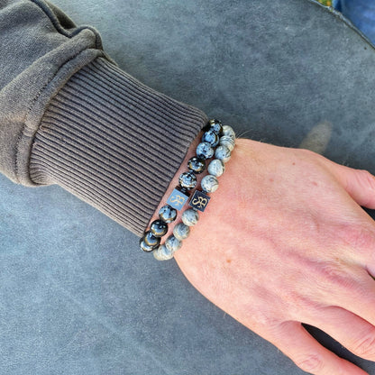 Een persoon die een donker shirt met lange mouwen draagt, toont zijn pols versierd met twee Stoney Bracelets' "Snowflake Obsidian Stones (8mm)" met zwart-witte gemarmerde patronen en vierkante metalen bedels. De hand rust op een grijs oppervlak.