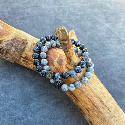 Twee kralenarmbanden van Stoney Bracelets rusten op een stuk rustiek hout. Elke armband toont een mix van glanzend zwart en gespikkelde grijsblauwe Snowflake Obsidian Stones (8 mm), met kleine metalen kubussen met symbolen verspreid tussen de kralen. De achtergrond is een getextureerd grijs oppervlak.