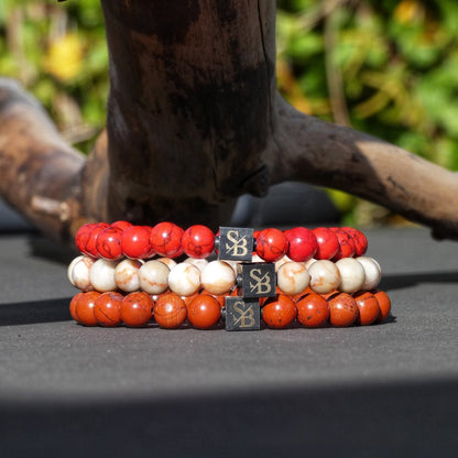 Drie kralen armbanden uit de Triple Red Set (8mm) van Stoney Bracelets worden op een oppervlak getoond. Deze armbanden zijn voorzien van natuursteen kralen in respectievelijk rood, wit en oranje, elk versierd met een gouden geaccentueerde zwarte kubus kraal. Op de achtergrond is een stuk drijfhout en wazig groen te zien.