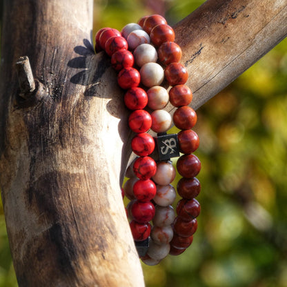 Vier Triple Red Set (8mm) kralenarmbanden van Stoney Bracelets zijn om een tak gewikkeld tegen een wazige groene en bruine achtergrond. Sommige kralen, gemaakt van natuursteen of edelsteen, tonen een logo met gestileerde letters.