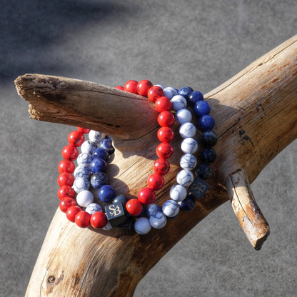 Drie Dutch Set (8mm) kralenarmbanden van Stoney Bracelets, gekleurd in de kleuren rood, blauw en wit met gemarmerde patronen, zijn gedrapeerd over een tak. Elke armband heeft een kleine zwarte vierkante kraal met een gouden logo erop. De achtergrond is wazig en grijs, waardoor de texturen en kleuren van de natuursteen kralen worden benadrukt.