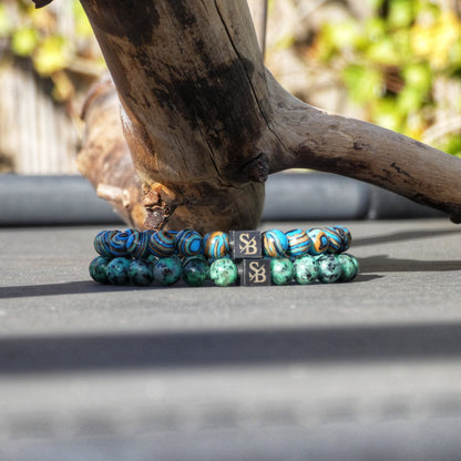 Twee Stoney Bracelets' Jupiter Mixed Stones (8mm) worden getoond, horizontaal op een grijs oppervlak. De armband bovenaan heeft blauwe, gele en zwarte wervelende edelsteen kralen, terwijl de armband eronder bestaat uit groene gemarmerde natuursteen kralen. Beide armbanden hebben kleine donkergrijze rechthoekige kralen met metalen "SB" logo's.