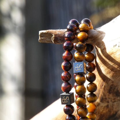 Close-up van twee kralenarmbanden uit de Tiger Eye Set (8mm) van Stoney Bracelets, één met donkerbruine natuursteen kralen en de andere met geelbruine edelsteen kralen, gedrapeerd over een houten tak. Beide armbanden zijn voorzien van vierkante metalen afstandhouders met gegraveerde symbolen.