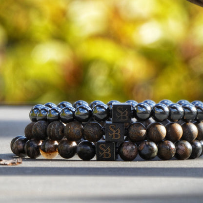 Er worden twee kralenarmbanden uit de Stoney Bracelets' Wood Set (8mm) collectie getoond, elk met donkerkleurige kralen. De ene armband is versierd met glanzende, zwarte kralen, terwijl de andere matte, donkerbruine kralen heeft. Beide armbanden hebben kleine vierkante bedeltjes gegraveerd met de letters "SB". De achtergrond is wazig.