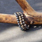 Een tak met een natuurlijke, gestructureerde look rust op een grijs oppervlak. Eromheen zitten twee Stoney Bracelets armbanden: een met metallic grijze kralen en de andere met de elegante Wood Set (8mm) versierd met houten kralen. De contrasterende kleuren en oppervlakteafwerkingen van de kralen benadrukken de schoonheid van de natuur.