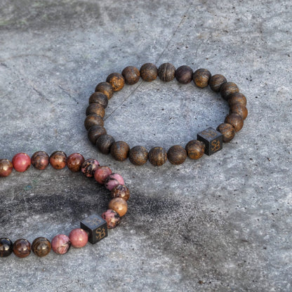 Op een getextureerd grijs oppervlak liggen twee armbanden van Stoney Bracelets. Eén armband toont gepolijste bruine Matte Bronziet Stones (8mm), terwijl de andere een mix van rode, bruine en roze natuursteen kralen bevat. Elke armband bevat een kleine zwarte kubus versierd met gouden initialen.