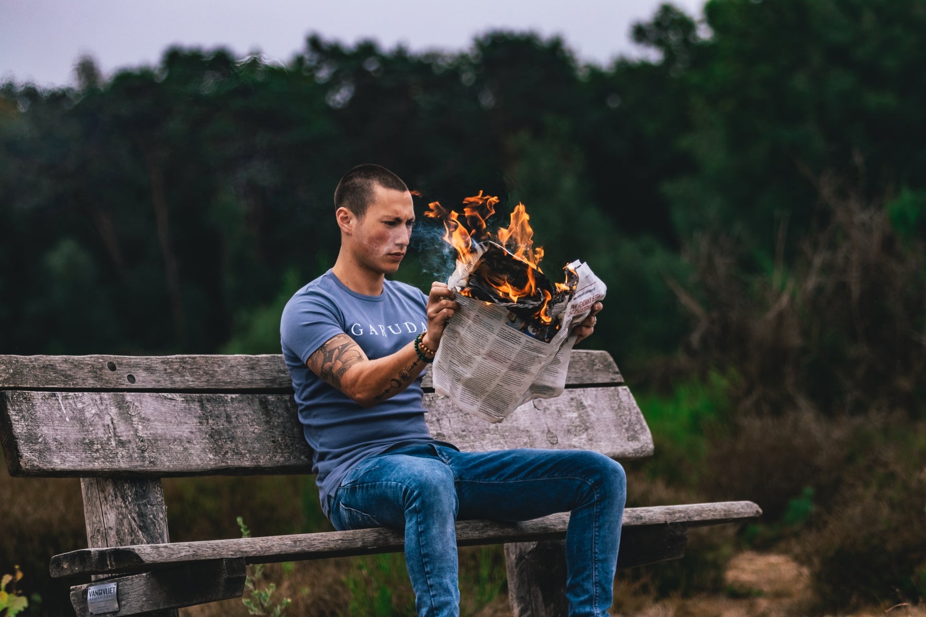 Een persoon zit buiten op een houten bankje en houdt een krant vast die in brand staat. Het individu, gekleed in een blauw shirt en jeans met een kralenarmband, lijkt naar de krant te kijken. Bomen en groen zijn zichtbaar op de achtergrond.