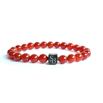 Een armband gemaakt van glanzende rode 8mm Red Onyx Stones, aan elkaar geregen met een kleine vierkante zilveren kraal met de letters "S" en "B" van Stoney Bracelets. De afbeelding heeft een witte achtergrond.