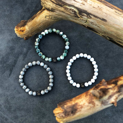 Drie Stoney-armbanden, met Picasso Jasper Stones (8 mm) in verschillende tinten en patronen, zijn in een driehoeksformatie op een donker oppervlak gerangschikt. Twee stukken verweerd hout omlijsten de boven- en rechterkant van de afbeelding. Elke armband heeft een klein vierkant label met een symbool.