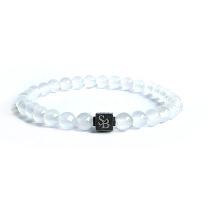 Een witte kralenarmband van Stoney Bracelets, met 6 mm witte onyxstenen en een zwarte vierkante bedel met de letters "SB". De armband, bestaande uit doorschijnende kralen die lijken op edelsteen, is gefotografeerd tegen een effen witte achtergrond.