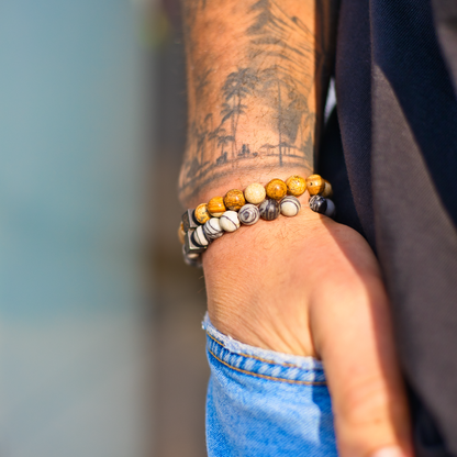 Een close-up van de pols van een persoon toont twee kralenarmbanden, een met Dessert Jasper Stones (8mm) van Stoney Bracelets met bruine kralen en de andere met zwart-witte kralenpatronen. De persoon draagt een blauwe spijkerbroek en heeft een tatoeage met boomontwerpen op zijn onderarm. Een armband heeft een edelsteen die prachtig het licht vangt.