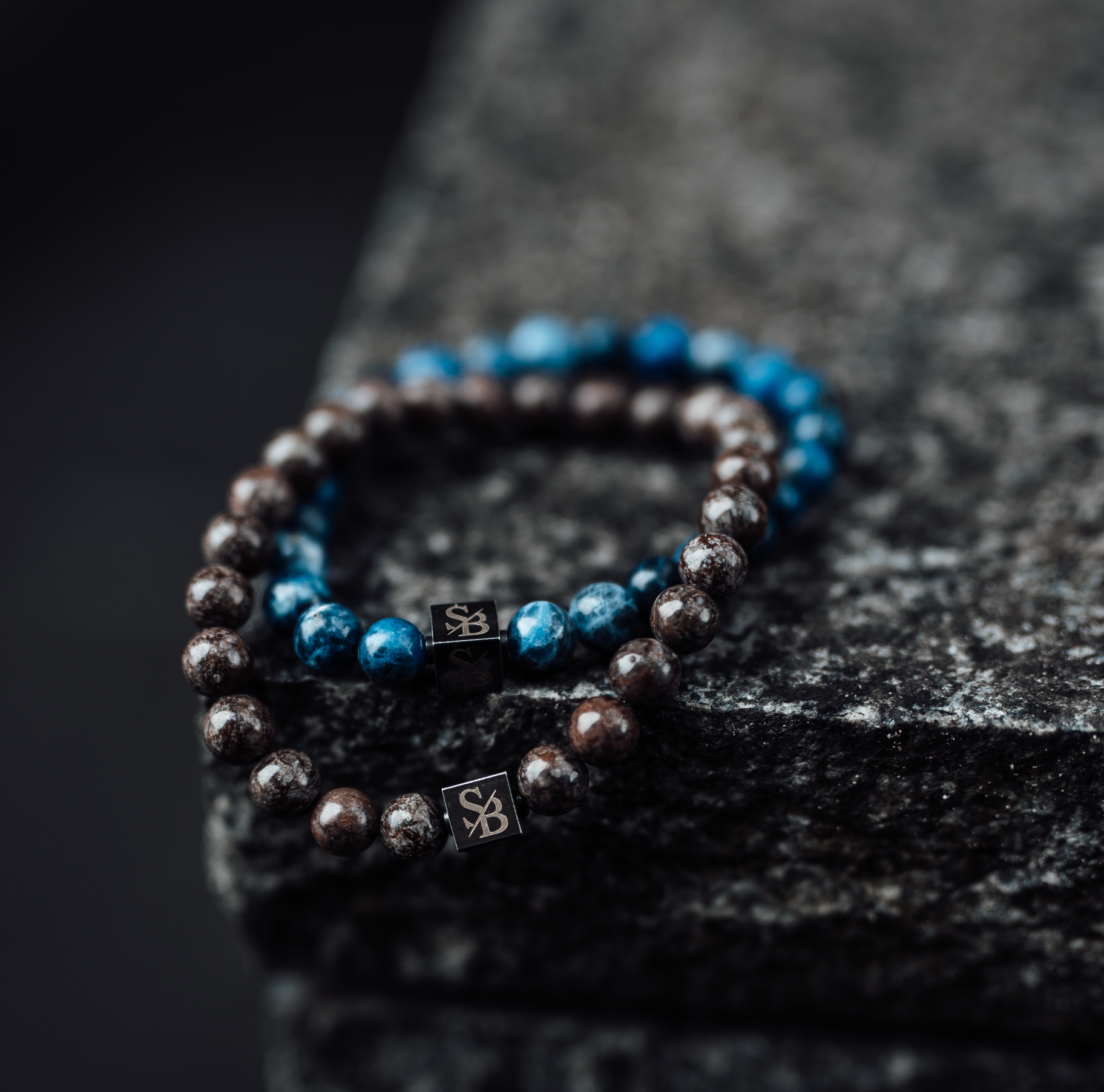 Twee kralen armbanden worden getoond op een getextureerd natuursteen oppervlak. De ene armband bestaat uit glanzende bruine kralen, en de andere heeft blauwe edelsteen kralen. Beide armbanden hebben een kleine rechthoekige metalen bedel met een "S B" gravure.