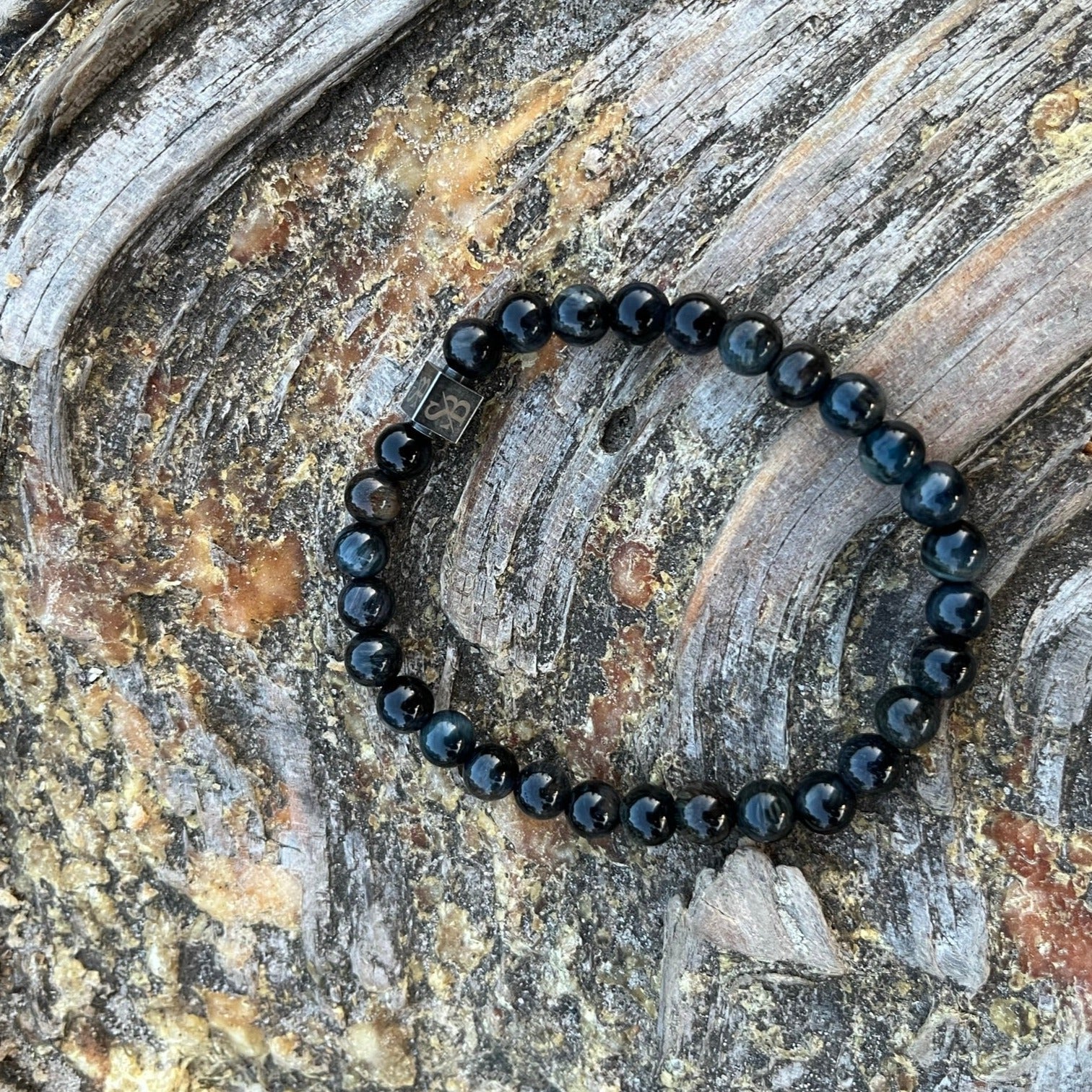 Een zwarte kralenarmband gemaakt van donkerblauwe Tijgeroogstenen (6 mm) zittend op een stuk hout van Stoney Bracelets.