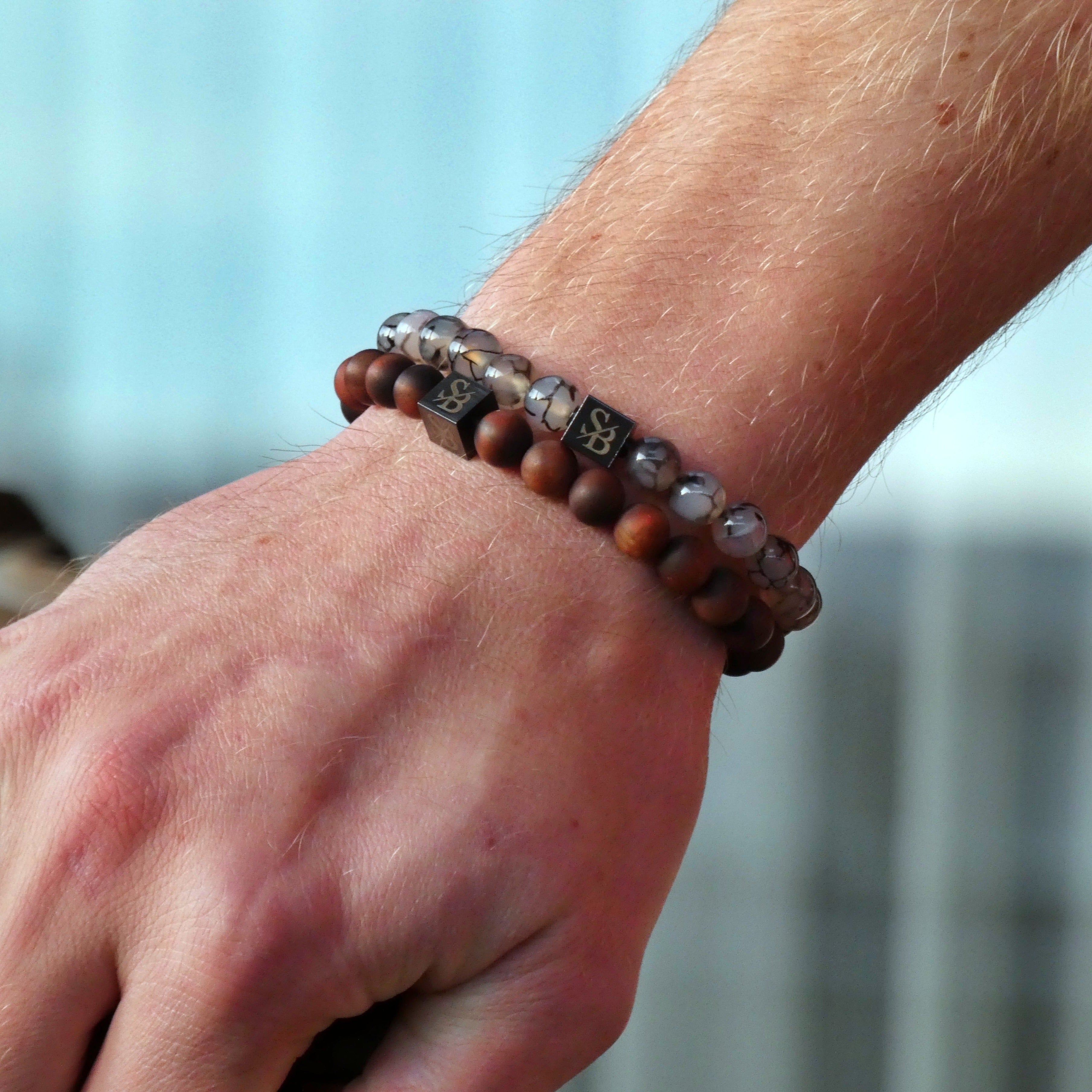 De hand van een man houdt een paar Stoney Bracelets Frosted Tiger Eye Stones (8 mm) armbanden vast.